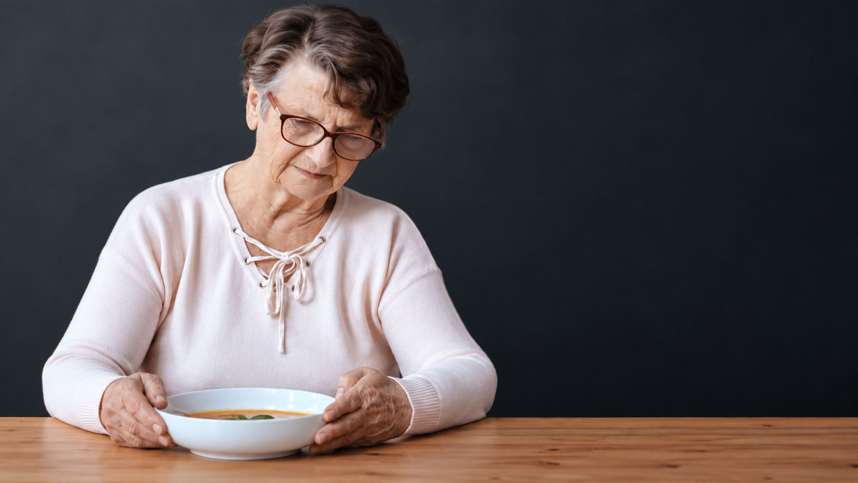 Older woman wearing glasses looking down at a bowl of food at a table.